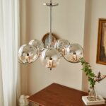 Silver-plated metal chandelier with 5 glass balls, arranged in a circle around a central axis. Installed in a room with beige walls, we can see the end of a white curtain on the left and a table with a book and a vase below the chandelier, as well as a piece of artwork on the right.