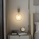 Wall-mounted lighted ball in a lighted circle. Hanging on the wall next to a bed, underneath a bedside table with an old-fashioned alarm clock resting on books.