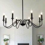 Black iron chandelier with 8 bulb candles. Simple and uncluttered. Installed in a living room. In the background is a white fireplace with a mirror and a plant in front of the mirror on either side of the fireplace.