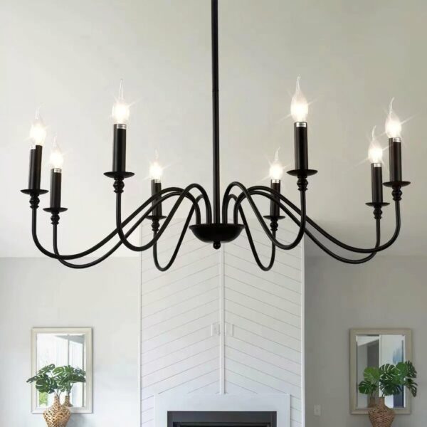 Black iron chandelier with 8 bulb candles. Simple and uncluttered. Installed in a living room. In the background is a white fireplace with a mirror and a plant in front of the mirror on either side of the fireplace.
