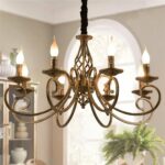 Gilded brass chandelier style with 6 heads with bulbs that replace the candle flames. The bulbs are lit. The chandelier hangs from the ceiling of a living room, with a green plant next to a window on the right and a piece of open furniture with knick-knacks and crockery on the left. The walls are beige.