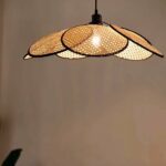 Cane chandelier in the shape of a flower, consisting of 6 mobile panels arranged around the lit bulb. Behind, a beige wall.