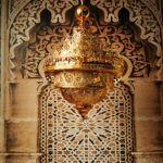 Large, majestic Moroccan gilded chandelier, with several crowns at the top giving the effect of flames, and the bottom descending to a point. The entire chandelier is hollowed out and perforated. Behind, a typical Moroccan wall with carved wood on one side and stone and tiles with Moroccan motifs on the other.