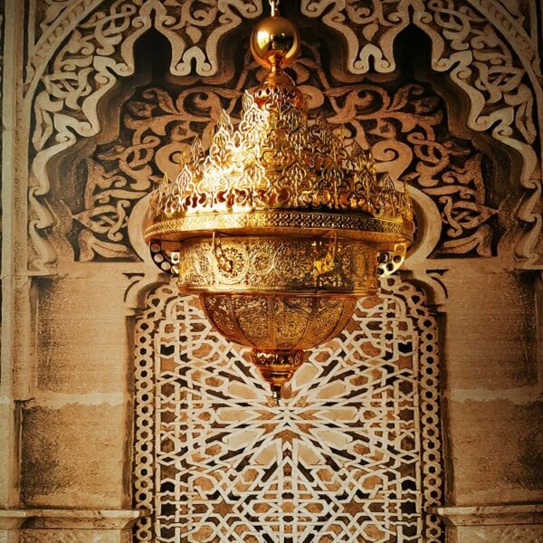 Large, majestic Moroccan gilded chandelier, with several crowns at the top giving the effect of flames, and the bottom descending to a point. The entire chandelier is hollowed out and perforated. Behind, a typical Moroccan wall with carved wood on one side and stone and tiles with Moroccan motifs on the other.