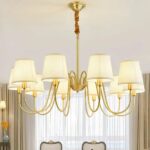 Gilded chandelier with 10 heads and white shades, suspended by a chain. We can see a window in the background and the backs of two chairs.