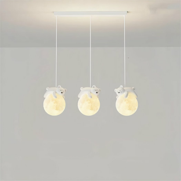 Hanging light, made up of three cables, each with a ball representing the moon, and a teddy bear sleeping on the moon. The balls are lit. The rectangular base hangs from the light-grey ceiling, with a light-grey wall in the background.