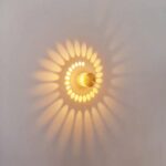 Wall light with effect, consisting of a grey metal cylinder, perforated with small holes, which lets the light spread in a spiral shape on the wall. On. Hung on a white wall.