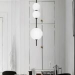 Vertical hanging chandelier made up of three lit white balls slid along a shaft. The lower ball is larger than the two smaller ones at the top. In the background, we see a white wall and white cupboard doors.