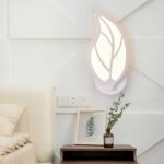 White wall light in the shape of a leaf, mounted on the wall and lit. It hangs on a white wall near a bed, above a dark-brown wooden bedside table. With a small green plant on top.