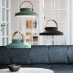 Three macaroon-shaped pendant lights with wooden handles hang above a gray sofa and a round white table. Bottom left, a light blue, above it offset to the left an olive green, and to the right a deep black. In the background is a white, checkered French window.