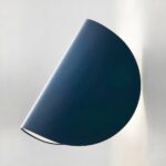 Blue wall sconce in the shape of a folded circle, towards the wall, like a taco. The bulb is lit and the wall is white.
