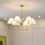 Gilded chandelier with copper body and 6 white fabric shades, accordion style. Hanging from a white ceiling, we see 3 spotlights in the background and a white wall with moldings.