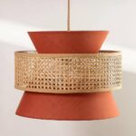 Terracotta and beige chandelier, suspended by a cable in the center. Composed of a terracotta-colored fabric shade in the shape of an hourglass, and a cane circle that surrounds the hourglass in the center. On a very light gray background.