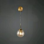 A modern wall sconce featuring a round gold base from which a slender gold stem extends downwards, at the end of which is a glass ball with a light bulb lit inside. On a black wall.