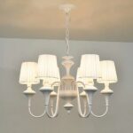 White baroque-style chandelier with 6 white shades, the bulbs are lit. Suspended by a chain from a white ceiling, a piece of white wall can be seen in the background.
