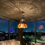Hand-woven straw and raffia chandelier, hung from the ceiling and lit, in a living-room-style room. Illuminates the ceiling with the play of shadows created by the weaving of raffia and straw.