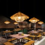 Natural straw chandelier, exotic straw hut style, hung on the wall and lit, above tables in what appears to be a restaurant, in a subdued lighting atmosphere.