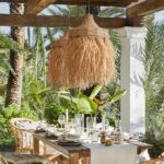 Hand-woven straw chandelier suspended under a pergola and above a dining table amid lush trees.