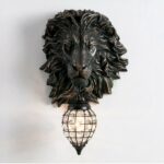 Lion-head wall sconce in black resin. Under its mouth, a drop-shaped lantern in black metal mesh. Imitation diamonds all around the lantern and a light bulb in the center.