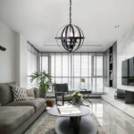 Black metal vintage chandelier in spherical form with 3 bulbs fixed inside the sphere like a candlestick. The ball is connected to the ceiling by a black chain. The chandelier is mounted in a living room above a brown oval coffee table, with an open magazine and a bouquet of white flowers. To the left, a gray sofa with a black-and-white striped cushion. Under the coffee table, a beige rug. On the wall opposite the sofa, a screen. Opposite, large windows with open Venetian blinds. In front of the windows, a green plant on the left and a brown armchair with beige cushions.