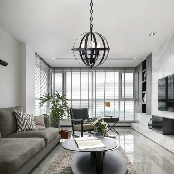 Black metal vintage chandelier in spherical form with 3 bulbs fixed inside the sphere like a candlestick. The ball is connected to the ceiling by a black chain. The chandelier is mounted in a living room above a brown oval coffee table, with an open magazine and a bouquet of white flowers. To the left, a gray sofa with a black-and-white striped cushion. Under the coffee table, a beige rug. On the wall opposite the sofa, a screen. Opposite, large windows with open Venetian blinds. In front of the windows, a green plant on the left and a brown armchair with beige cushions.